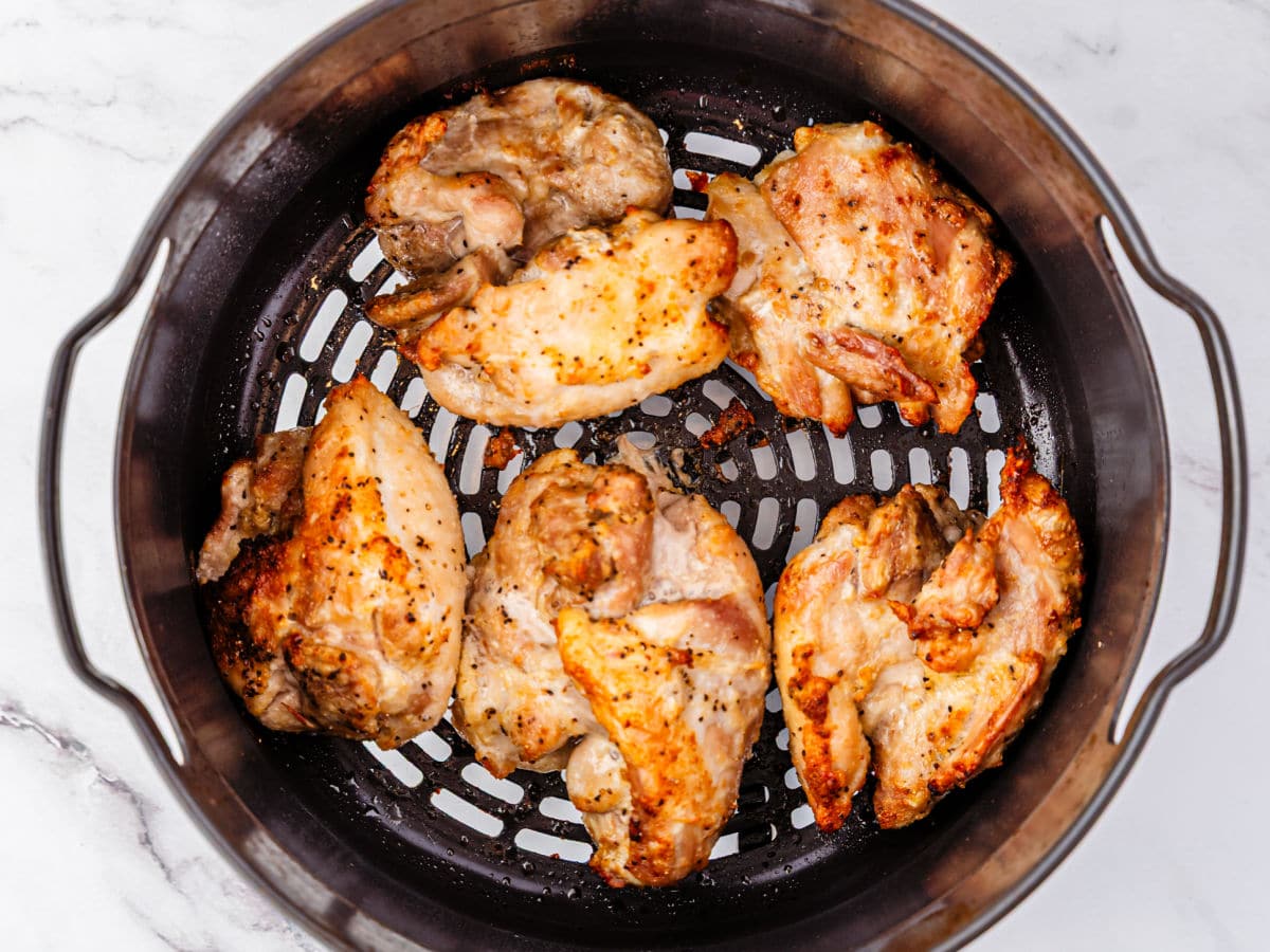 Six pieces of cooked, seasoned Garlic Parmesan Chicken are arranged in a round air fryer basket with a black perforated base, seen from above on a light marble surface.