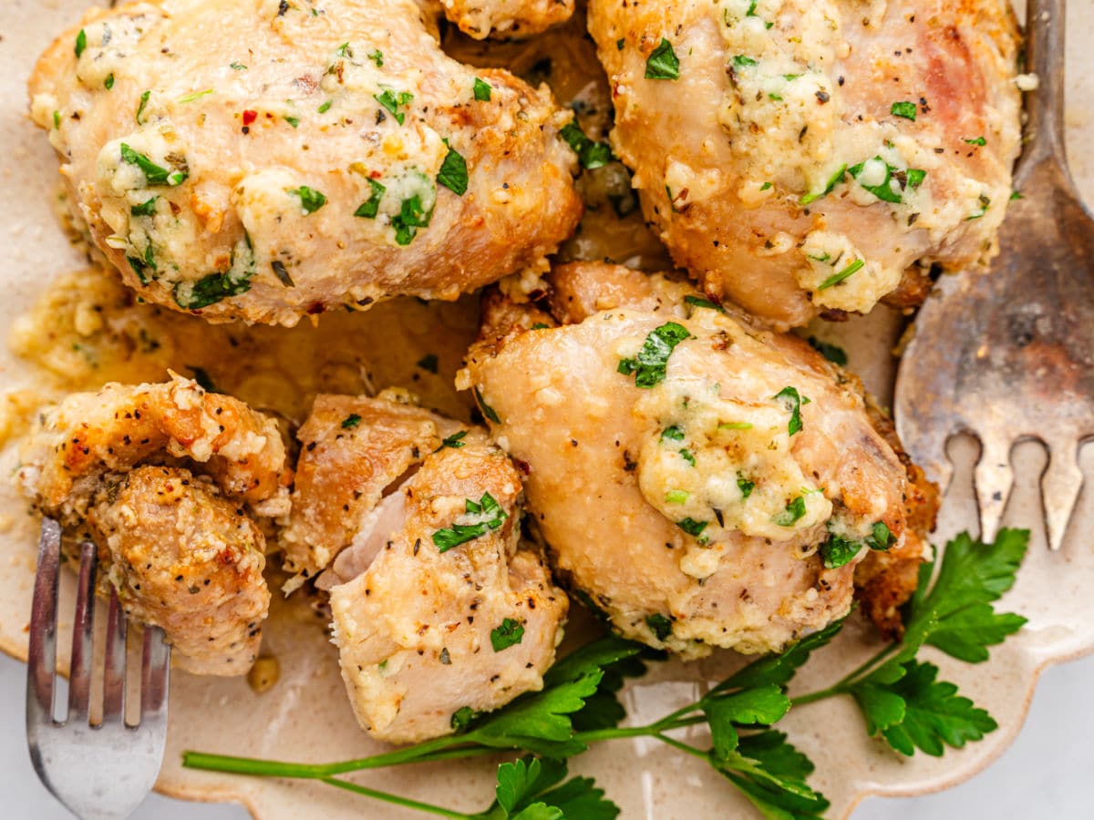 Close-up of Garlic Parmesan Chicken Air Fryer pieces in a creamy garlic sauce, garnished with chopped parsley on a beige plate. A fork and serving spoon rest nearby, with fresh parsley leaves beside the chicken.