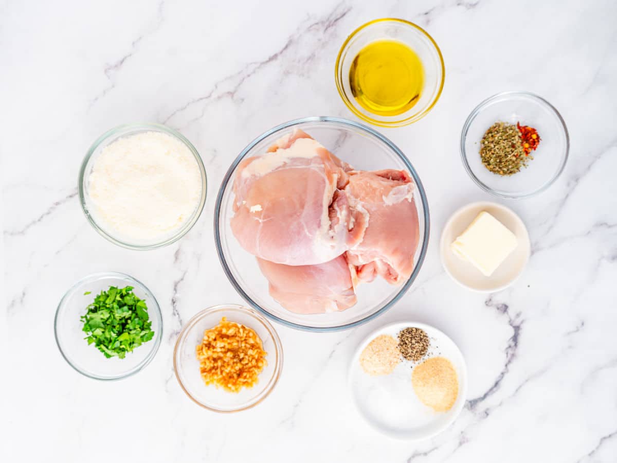 A top view of raw chicken thighs in a bowl surrounded by small bowls of grated cheese, olive oil, chopped parsley, minced garlic, butter, spices, and seasonings—perfect for preparing Garlic Parmesan Chicken Air Fryer style on a white marble surface.