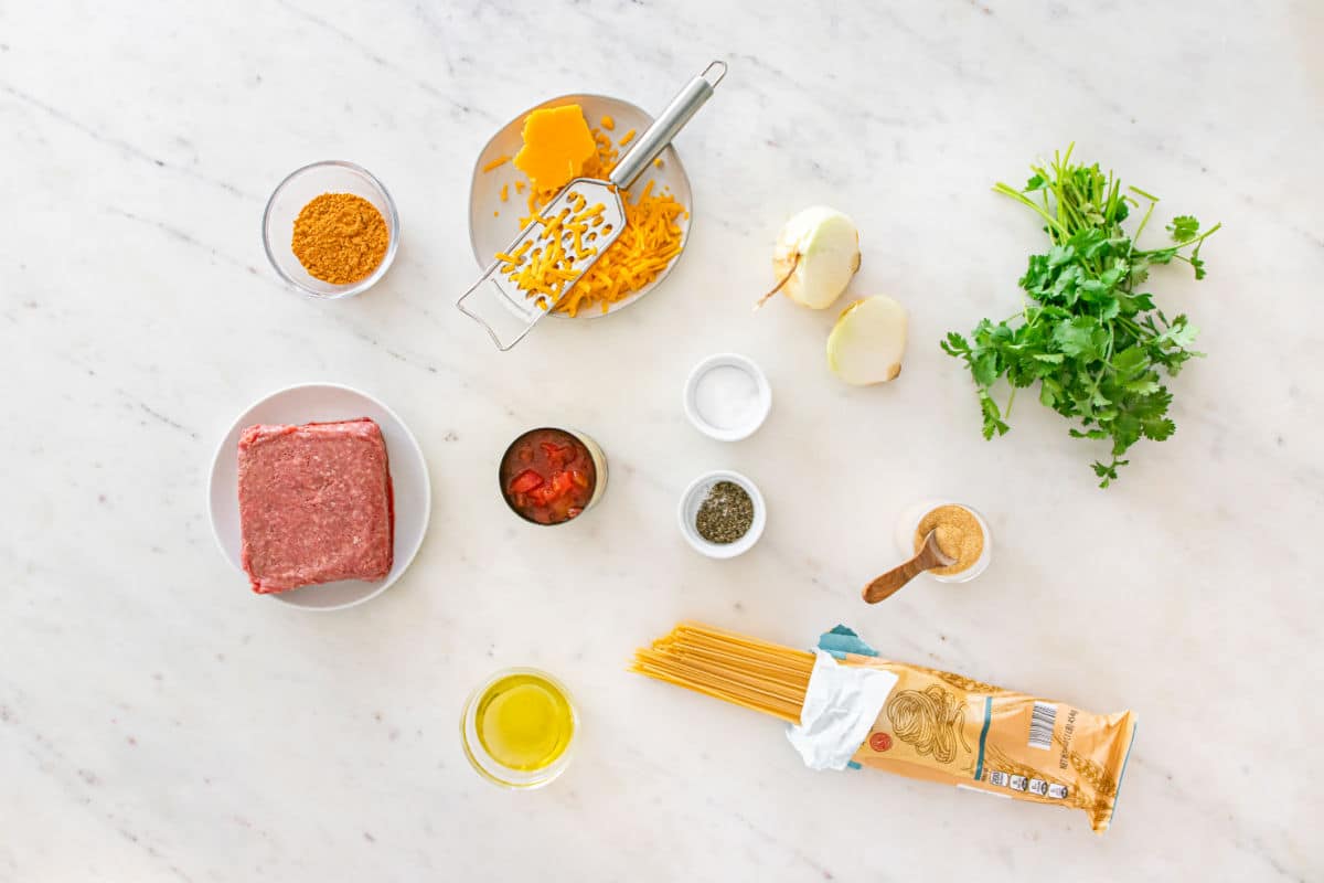Ingredients for taco spaghetti arranged on a white surface: ground beef, dry spaghetti, olive oil, chopped cheese, onion, fresh cilantro, canned tomatoes, grated cheese, and various spices in small bowls.