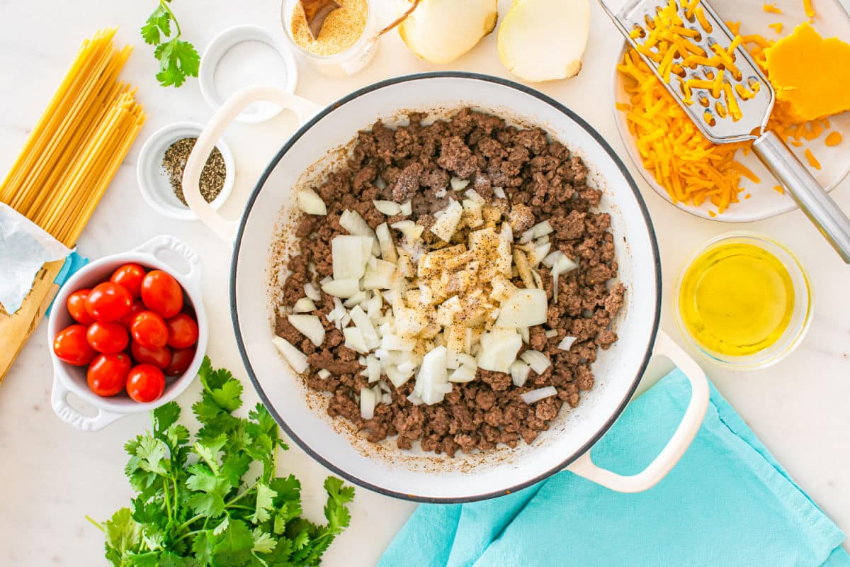 A white pot filled with ground beef, chopped onions, and seasonings sits on a counter surrounded by uncooked spaghetti, cherry tomatoes, cilantro, shredded cheese, oil, sliced onions, and bowls of spices—perfect for making taco spaghetti.