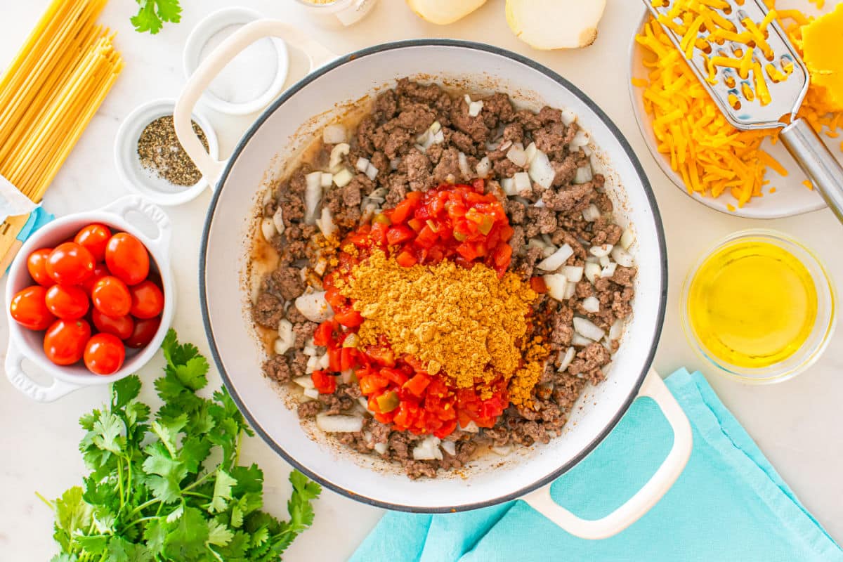 Enamel pot filled with cooked ground beef, chopped onions, diced tomatoes, and taco seasoning for a delicious taco spaghetti. Surrounding are cherry tomatoes, shredded cheese, oil, cilantro, spaghetti, seasonings, and a turquoise towel.