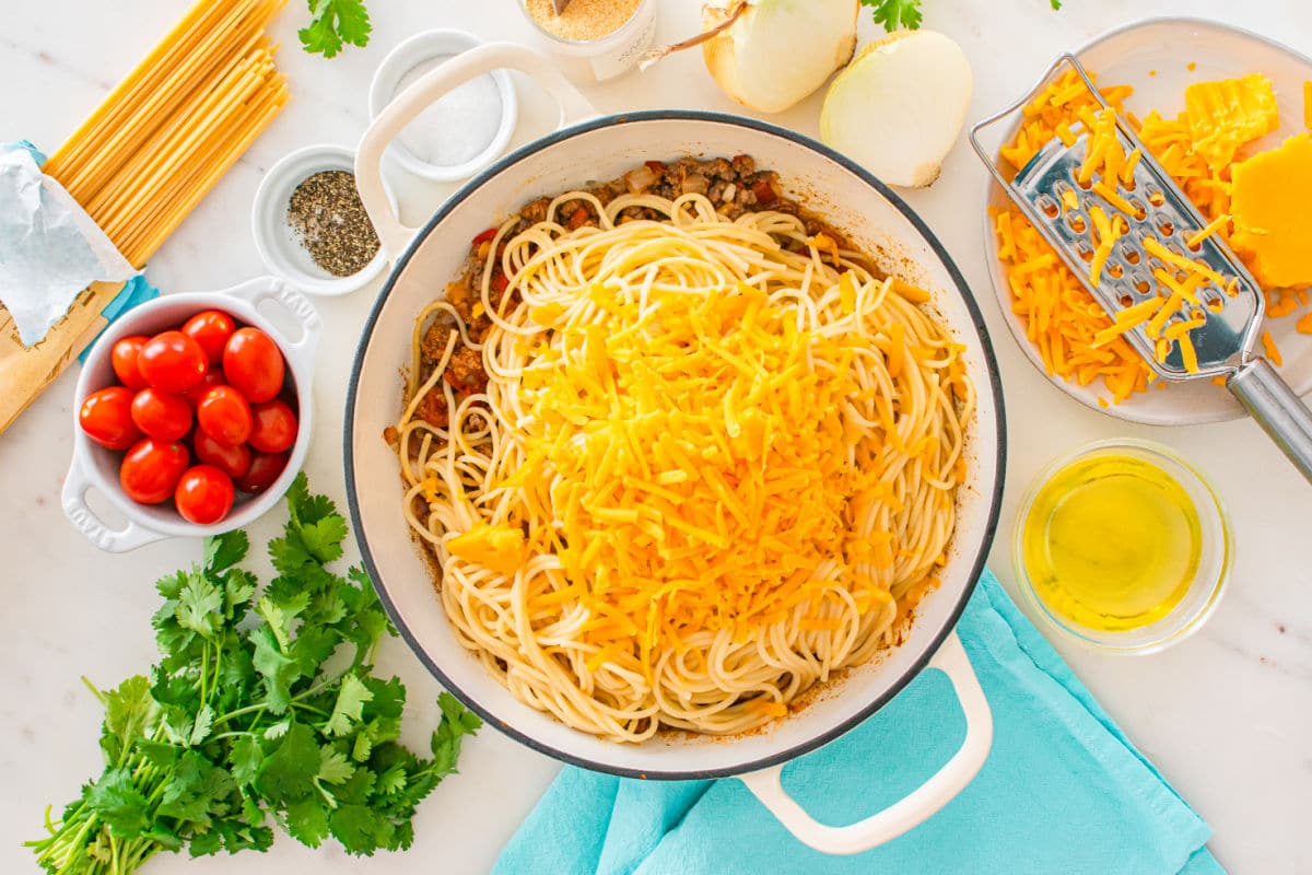 A pot of cooked taco spaghetti topped with shredded cheddar cheese sits on a counter surrounded by cherry tomatoes, cilantro, oil, chopped onions, black pepper, dry spaghetti, and a cheese grater.