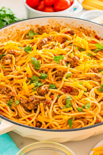 A skillet filled with taco spaghetti—spaghetti mixed with ground beef, tomato sauce, and topped with shredded cheddar cheese and fresh parsley. Fresh ingredients and kitchen utensils are visible in the background.