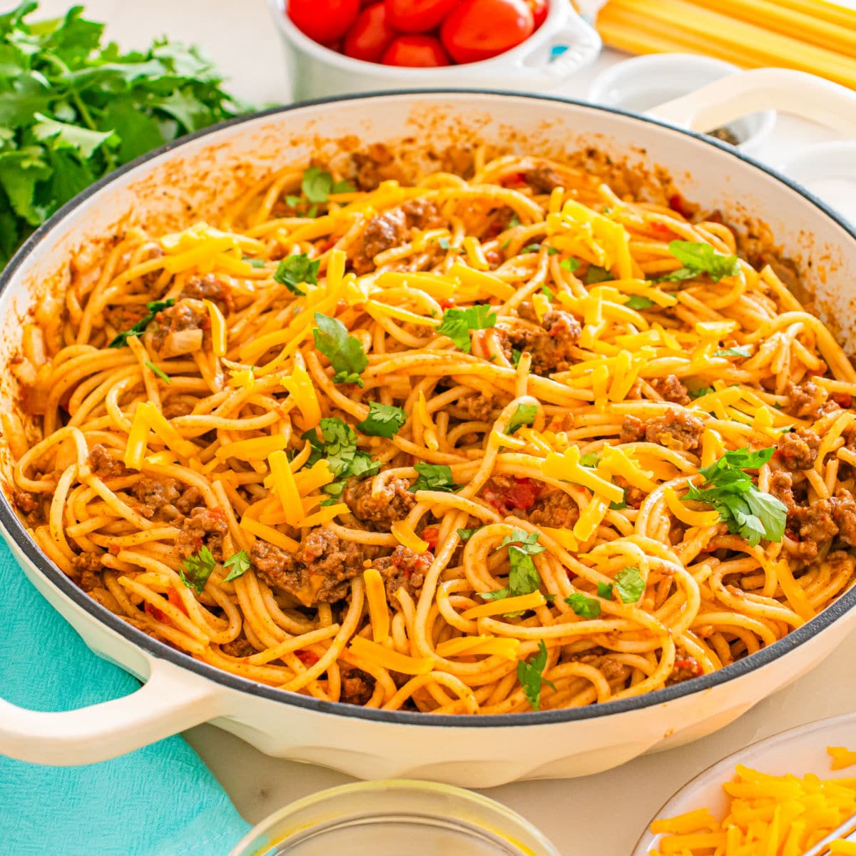 A skillet filled with taco spaghetti—spaghetti mixed with ground beef, tomato sauce, and topped with shredded cheddar cheese and fresh parsley. Fresh ingredients and kitchen utensils are visible in the background.