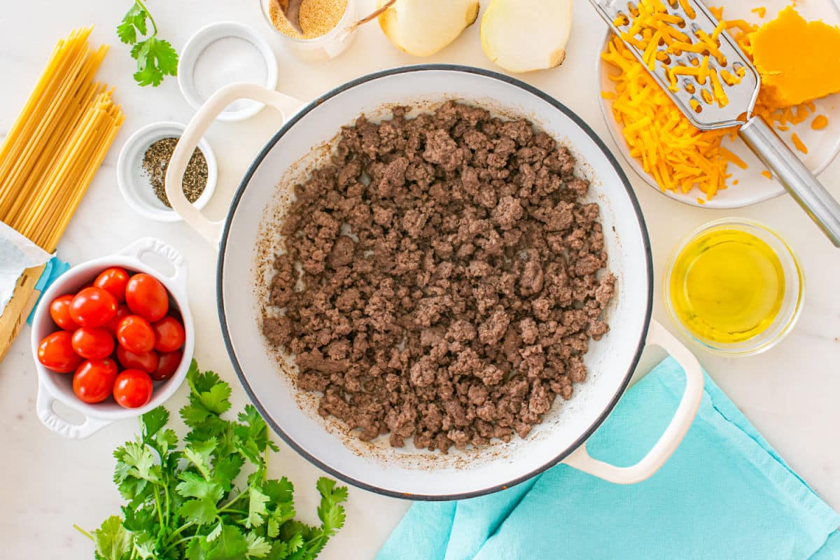 A white pot filled with cooked ground beef for taco spaghetti is surrounded by ingredients including spaghetti, cherry tomatoes, cilantro, shredded cheddar cheese, onions, olive oil, and seasonings on a white countertop.