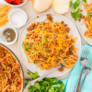A table set with plates of taco spaghetti topped with ground meat, shredded cheese, and fresh herbs, surrounded by uncooked pasta, onions, cilantro, and seasonings. Forks and a blue napkin are placed nearby.