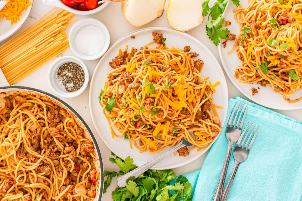 A table set with plates of taco spaghetti topped with ground meat, shredded cheese, and fresh herbs, surrounded by uncooked pasta, onions, cilantro, and seasonings. Forks and a blue napkin are placed nearby.