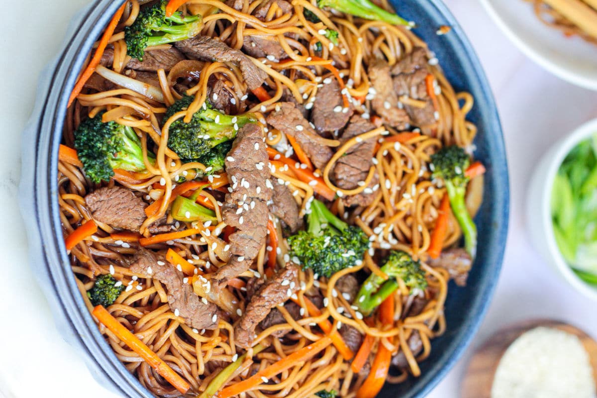 A bowl of Beef Noodle Stir Fry featuring sliced beef, broccoli, carrots, and sesame seeds, all tossed in a savory sauce and served in a blue dish.