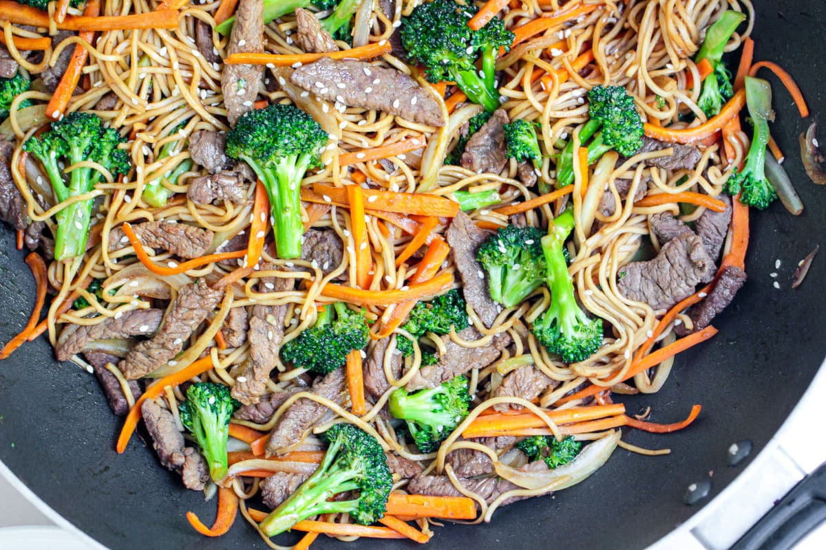 A close-up of a Beef Noodle Stir Fry in a pan, featuring noodles, slices of beef, broccoli florets, carrot strips, onion, and sesame seeds, all mixed together.