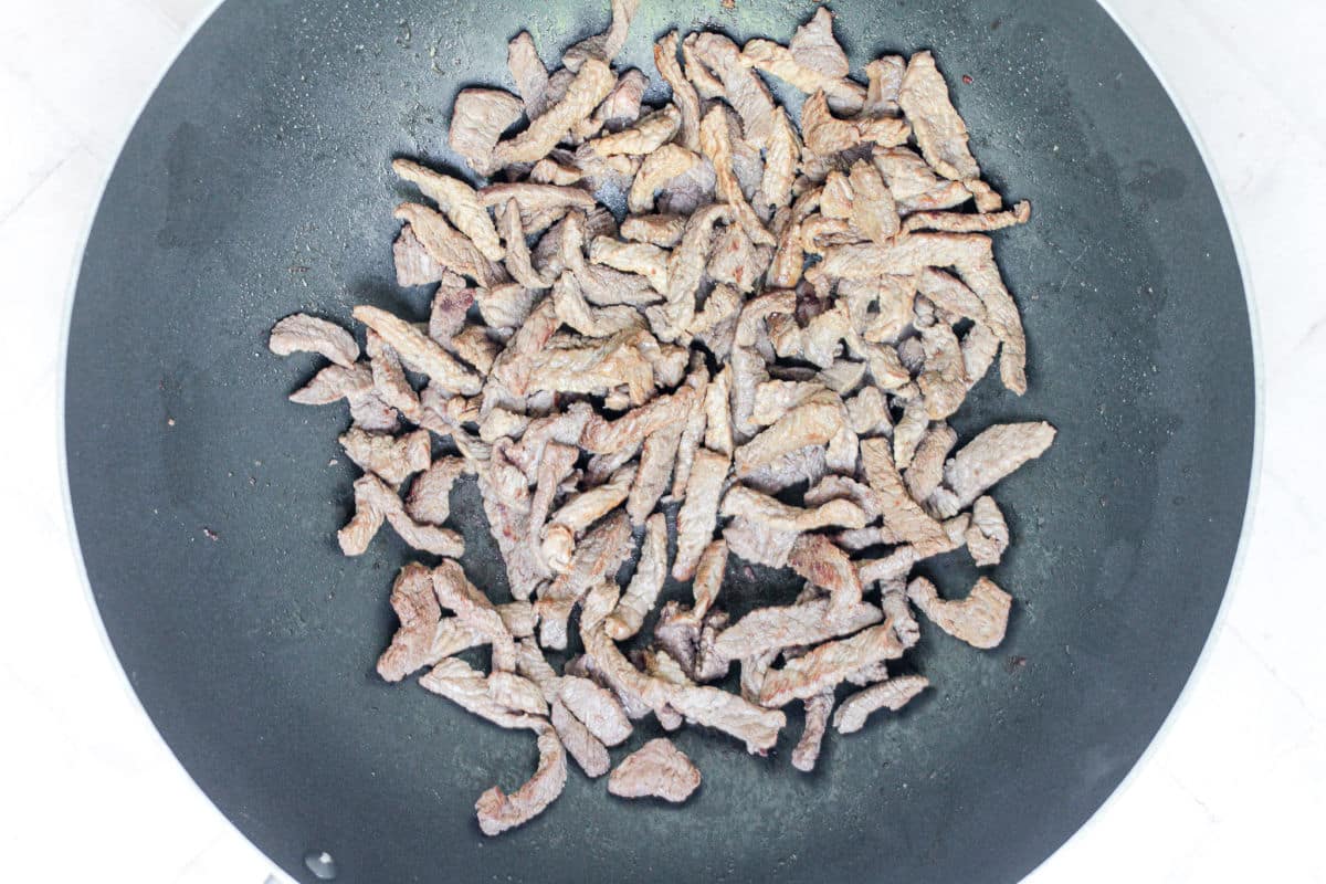 Thin strips of beef for Beef Noodle Stir Fry sizzling and browning in a large nonstick skillet on a stove, viewed from above.