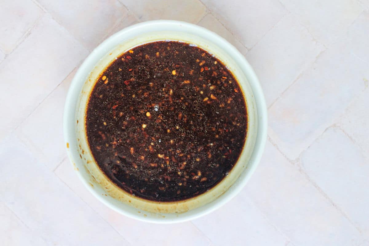 A white bowl filled with a dark, speckled sauce containing visible chili flakes and seasonings, perfect for drizzling over Beef Noodle Stir Fry, placed on a light tiled surface.