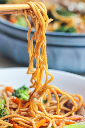 Chopsticks lifting a portion of Beef Noodle Stir Fry with vegetables, including broccoli and carrots, from a white bowl, with a pan of more noodles visible in the background.