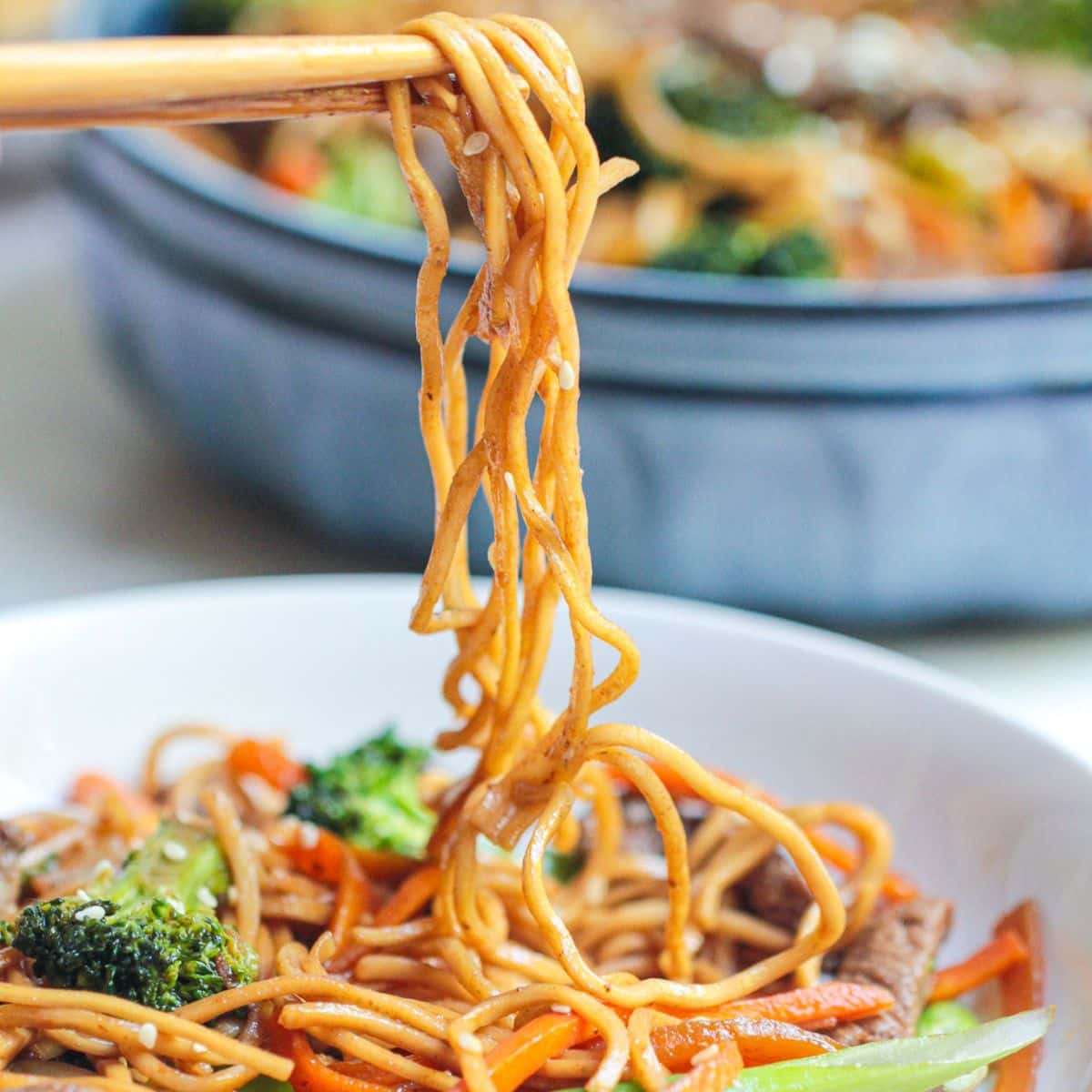 Chopsticks lifting a portion of Beef Noodle Stir Fry with vegetables, including broccoli and carrots, from a white bowl, with a pan of more noodles visible in the background.