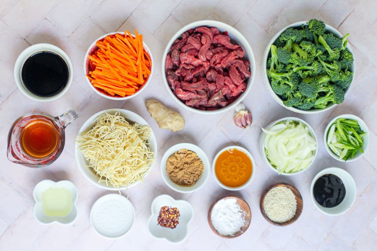 An overhead view of bowls holding Beef Noodle Stir Fry ingredients—sliced beef, broccoli, carrot strips, noodles, sliced onion, green onions, garlic, ginger, and various sauces and seasonings—all arranged neatly on a light-colored surface.
