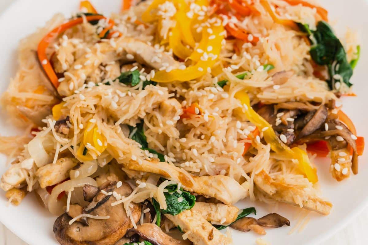 A plate of stir-fried rice noodles with sliced mushrooms, bell peppers, spinach, tofu strips, and sesame seeds, served on a white dish.
