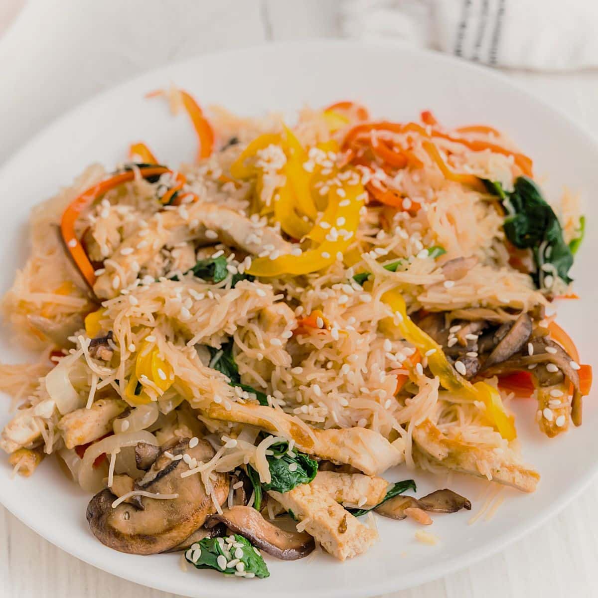 A white plate filled with Tofu Japchae with Vermicelli—stir-fried rice noodles, sliced bell peppers, mushrooms, spinach, and strips of tofu, all topped with sesame seeds.
