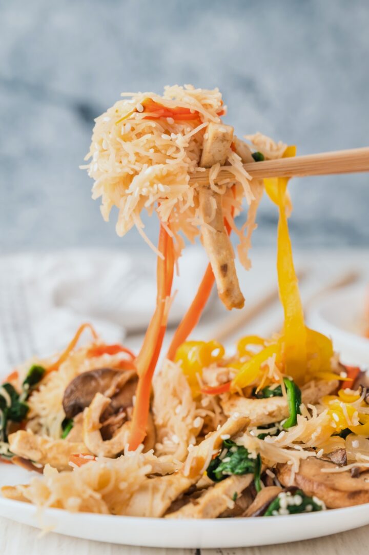 Chopsticks lifting noodles, vegetables, and tofu from a plate of stir-fried noodles with colorful bell peppers, carrots, mushrooms, and sesame seeds.