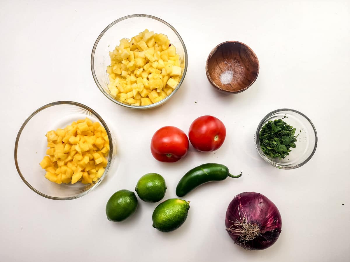 Chopped yellow and orange mango, diced pineapple, salt, two tomatoes, chopped cilantro, three limes, a jalapeño pepper, and red onion arranged on a white surface—everything you need for fresh Mango Pineapple Salsa.