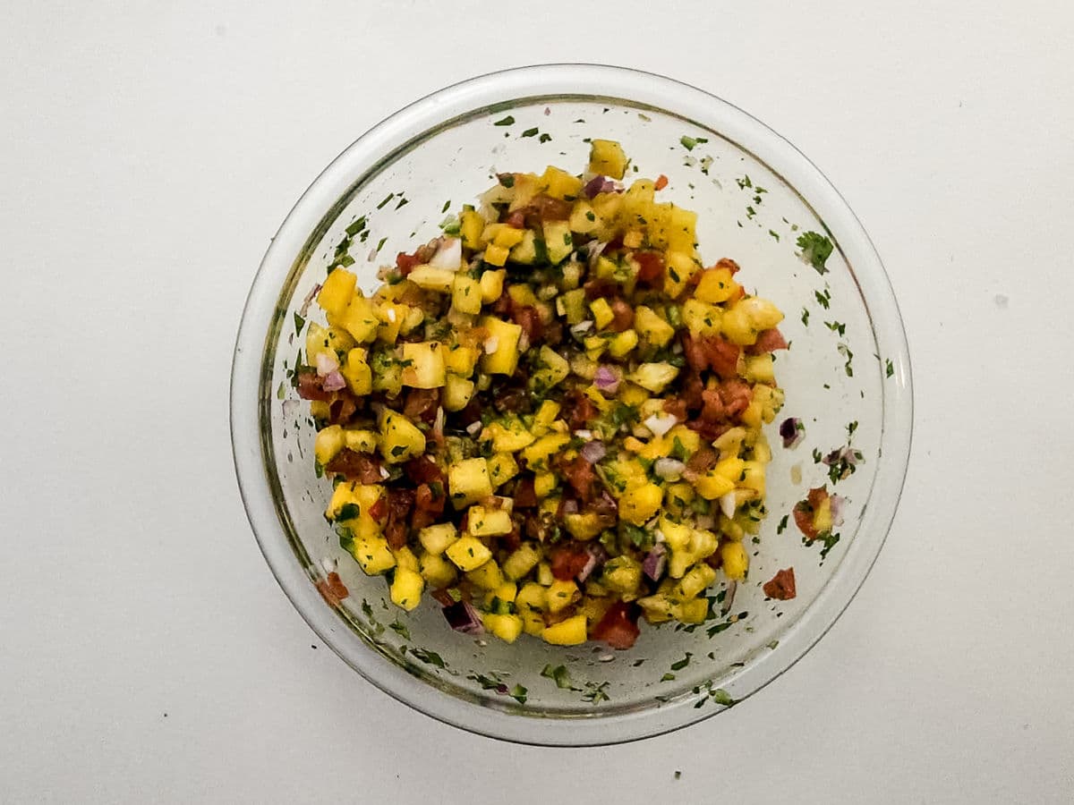 A glass bowl filled with vibrant Mango Pineapple Salsa—diced mango, pineapple, tomatoes, red onion, cilantro, and other fresh ingredients—sits on a plain white surface.