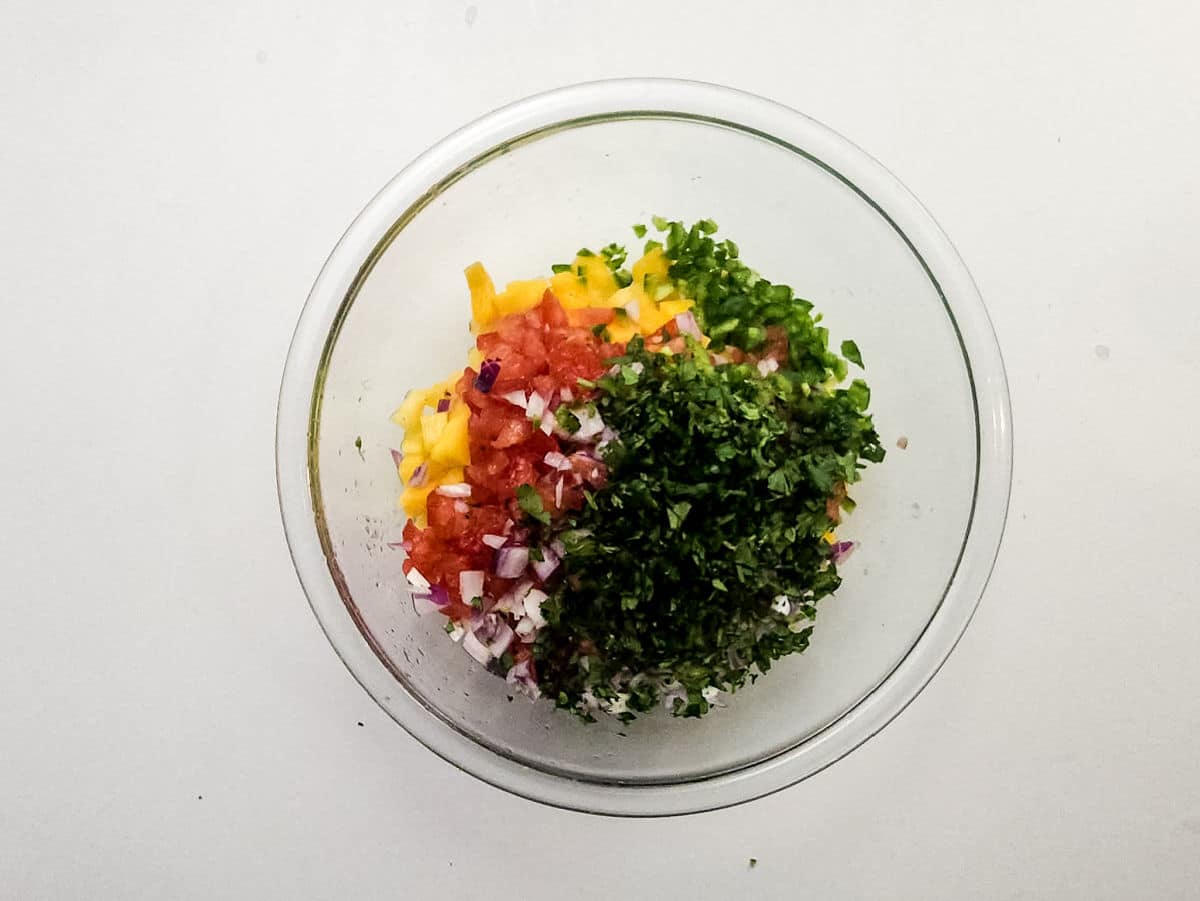 A glass bowl on a white surface contains chopped yellow bell pepper, tomato, red onion, green jalapeño, and fresh cilantro—colorful ingredients perfectly arranged and ready to be mixed for a vibrant Mango Pineapple Salsa.