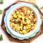 A bowl of Mango Pineapple Salsa with diced pineapple, mango, tomatoes, onions, cilantro, and lime in a white bowl on stacked blue and wooden plates, with tortilla chips and cilantro leaves nearby.