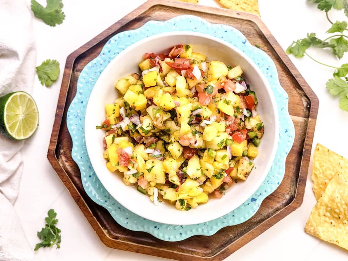 A bowl of Mango Pineapple Salsa with diced pineapple, mango, tomatoes, onions, cilantro, and lime in a white bowl on stacked blue and wooden plates, with tortilla chips and cilantro leaves nearby.