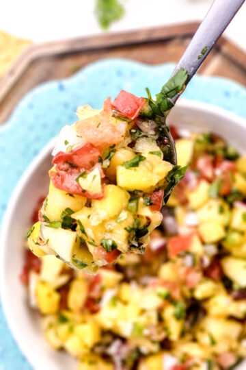 A close-up of a spoonful of Mango Pineapple Salsa with diced tomatoes, avocado, mango, cilantro, and onion, held over a bowl of the same salsa. Tortilla chips are visible in the background.