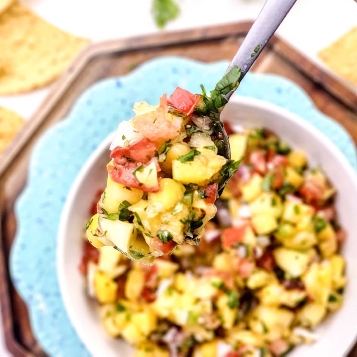 A close-up of a spoonful of Mango Pineapple Salsa with diced tomatoes, avocado, mango, cilantro, and onion, held over a bowl of the same salsa. Tortilla chips are visible in the background.