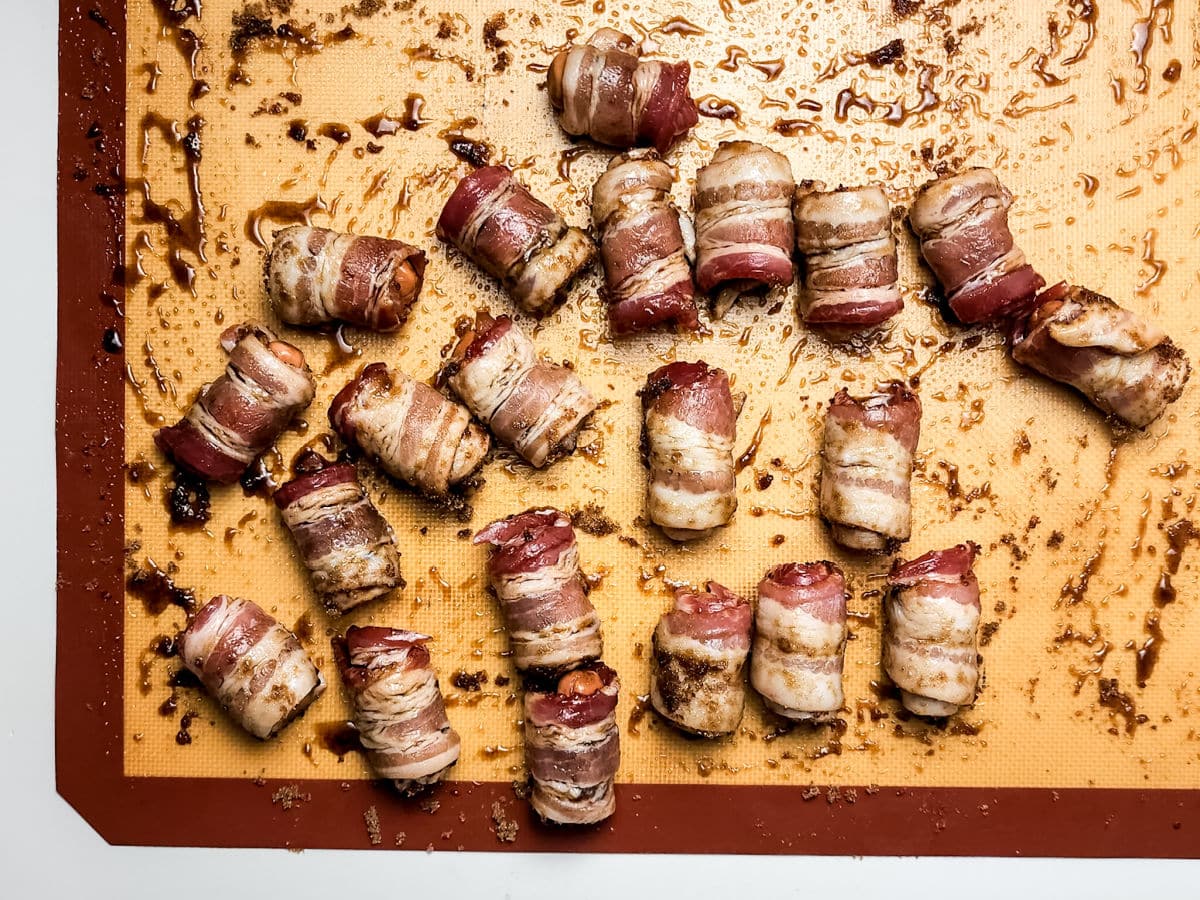 Maple bacon smokies-wrapped dates arranged on a silicone baking mat, freshly baked with caramelized bits and juices scattered around.