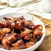 A white bowl filled with maple bacon smokies, each wrapped in bacon and secured with a toothpick, sits on a wooden board. The sausages are glazed and appear caramelized.
