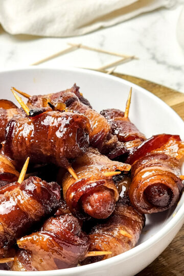 A white bowl filled with maple bacon smokies, each wrapped in bacon and secured with a toothpick, sits on a wooden board. The sausages are glazed and appear caramelized.
