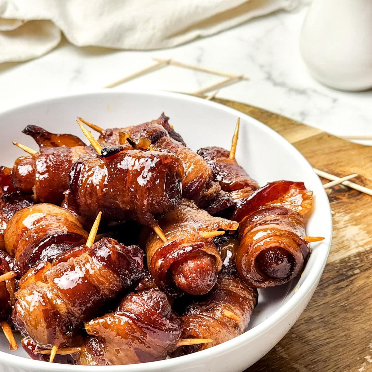 A white bowl filled with maple bacon smokies, each wrapped in bacon and secured with a toothpick, sits on a wooden board. The sausages are glazed and appear caramelized.