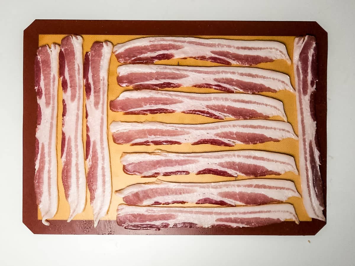 Raw bacon strips are neatly arranged in rows on a brown cutting board with a yellow surface, ready to be used for maple bacon smokies, set against a plain white background.