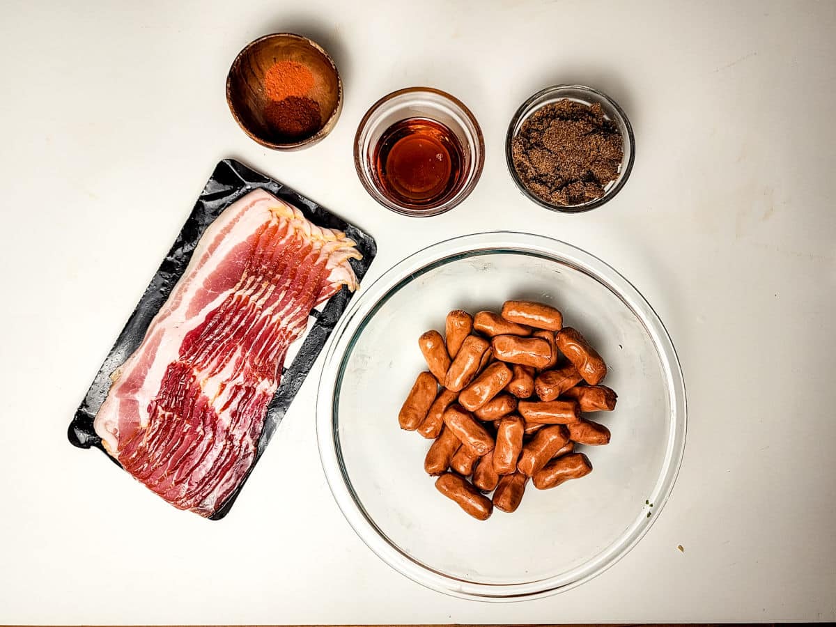 Overhead view of slices of raw bacon, a bowl of cocktail sausages for maple bacon smokies, and three small bowls containing paprika, maple syrup, and brown sugar on a white surface.