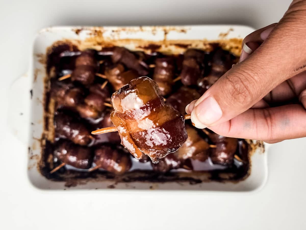 A hand holds a toothpick with a bacon-wrapped date above a baking dish filled with more maple bacon smokies, all glazed and perfectly caramelized.