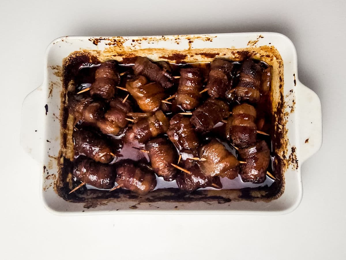 A white rectangular baking dish filled with maple bacon smokies—bacon-wrapped dates secured with toothpicks, baked in a dark, sticky sauce. The dish shows caramelized edges and cooked bacon rolls nestled in the rich sauce.
