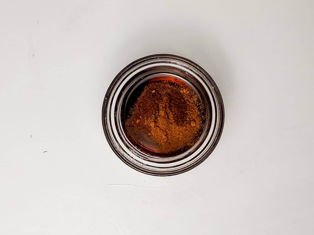 A small glass bowl filled with a reddish-brown powder, possibly a spice or seasoning for maple bacon smokies, sits on a plain white surface.