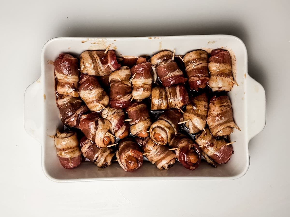 A white casserole dish filled with maple bacon smokies—bacon-wrapped sausages secured with toothpicks, arranged in rows and baked until browned.