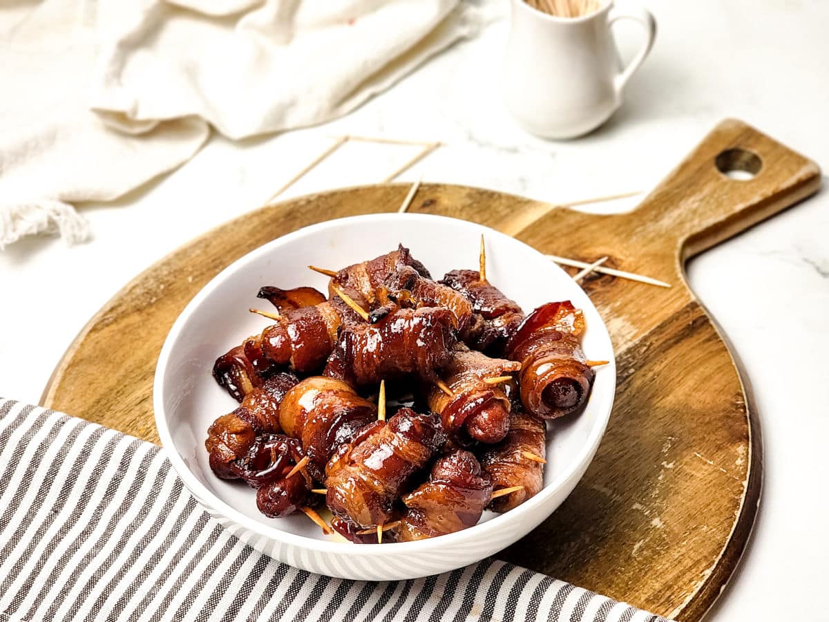A white bowl filled with maple bacon smokies, some secured with toothpicks, sits on a wooden cutting board next to a striped cloth and a small white pitcher.
