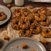 A pile of Somoa Copy Cat Cookies, caramel and chocolate-drizzled, sits on parchment paper surrounded by shredded coconut, caramel squares, plates of cookies, a bowl of toasted coconut, and two glass bottles of milk.
