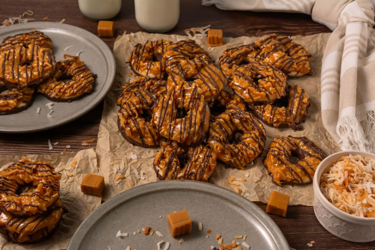 A pile of Somoa Copy Cat Cookies, caramel and chocolate-drizzled, sits on parchment paper surrounded by shredded coconut, caramel squares, plates of cookies, a bowl of toasted coconut, and two glass bottles of milk.