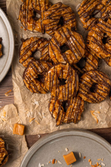 A group of Somoa Copy Cat Cookies, caramel and chocolate-drizzled with coconut flakes, is arranged on parchment paper and gray plates, surrounded by caramel cubes and coconut shavings on a wooden surface.