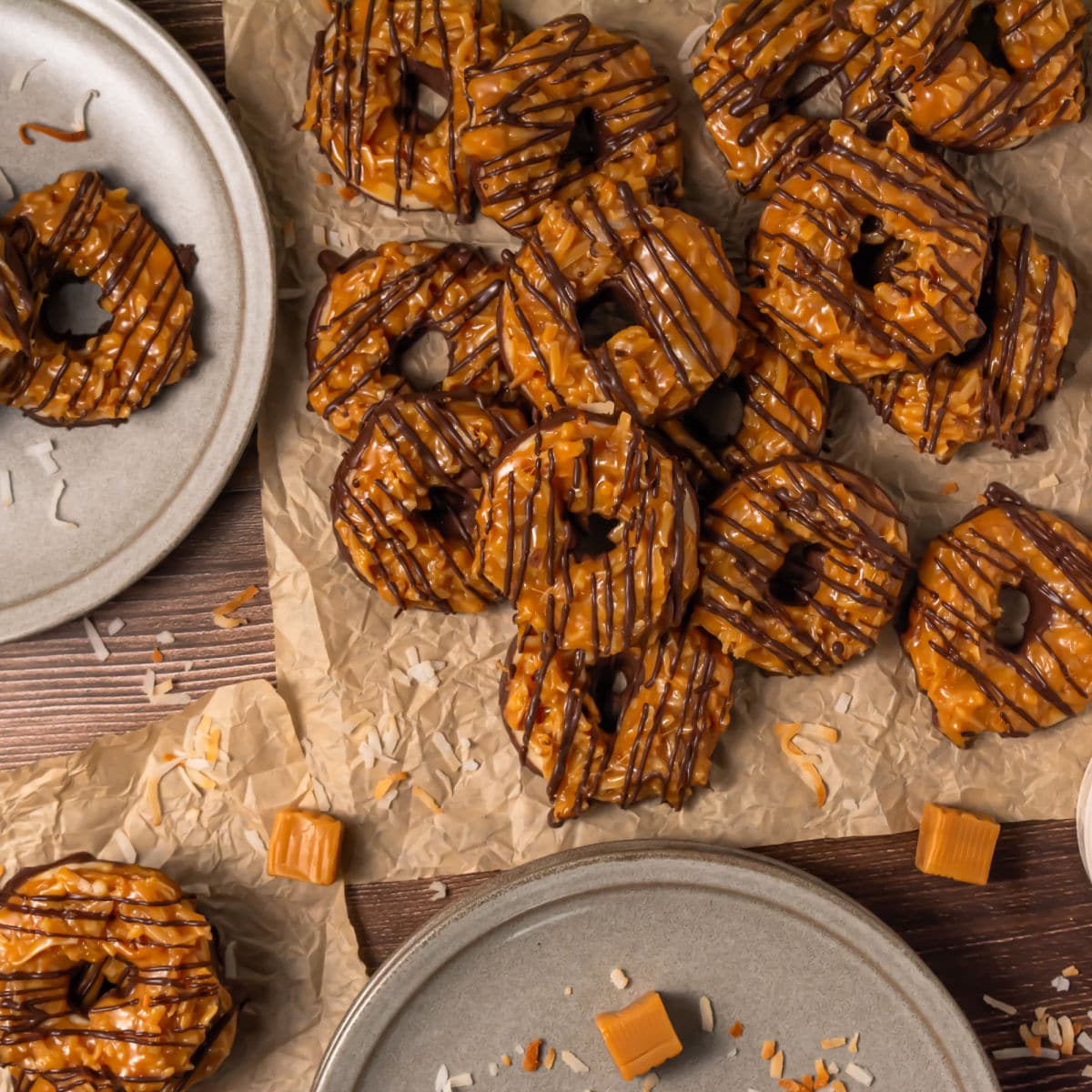 A group of Somoa Copy Cat Cookies, caramel and chocolate-drizzled with coconut flakes, is arranged on parchment paper and gray plates, surrounded by caramel cubes and coconut shavings on a wooden surface.