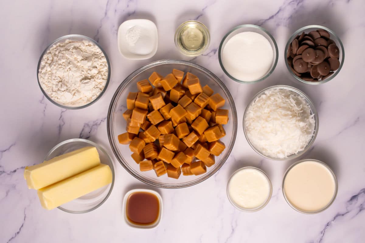 Various baking ingredients are arranged in bowls on a marble surface, ready for Somoa Copy Cat Cookies, including flour, sugar, chocolate, coconut, caramel candies, butter, vanilla extract, cream, condensed milk, oil, and baking powder.