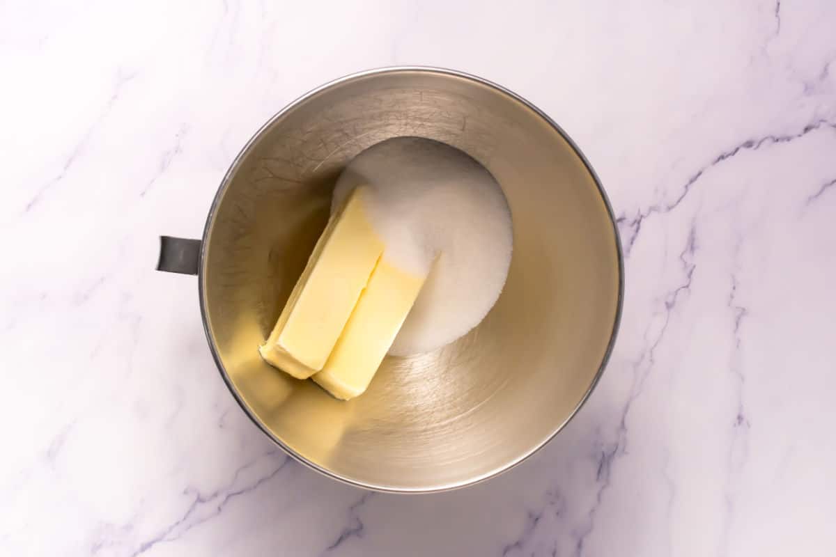 A metal mixing bowl containing two sticks of butter and a pile of granulated sugar, viewed from above on a white marble surface, ready to create the perfect Somoa Copy Cat Cookies.