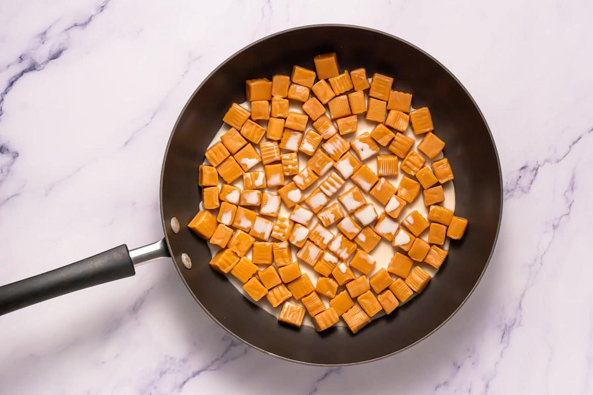 A nonstick frying pan with unwrapped caramel candies and a small amount of milk inside, perfect for making Somoa Copy Cat Cookies, sits on a white marble surface.