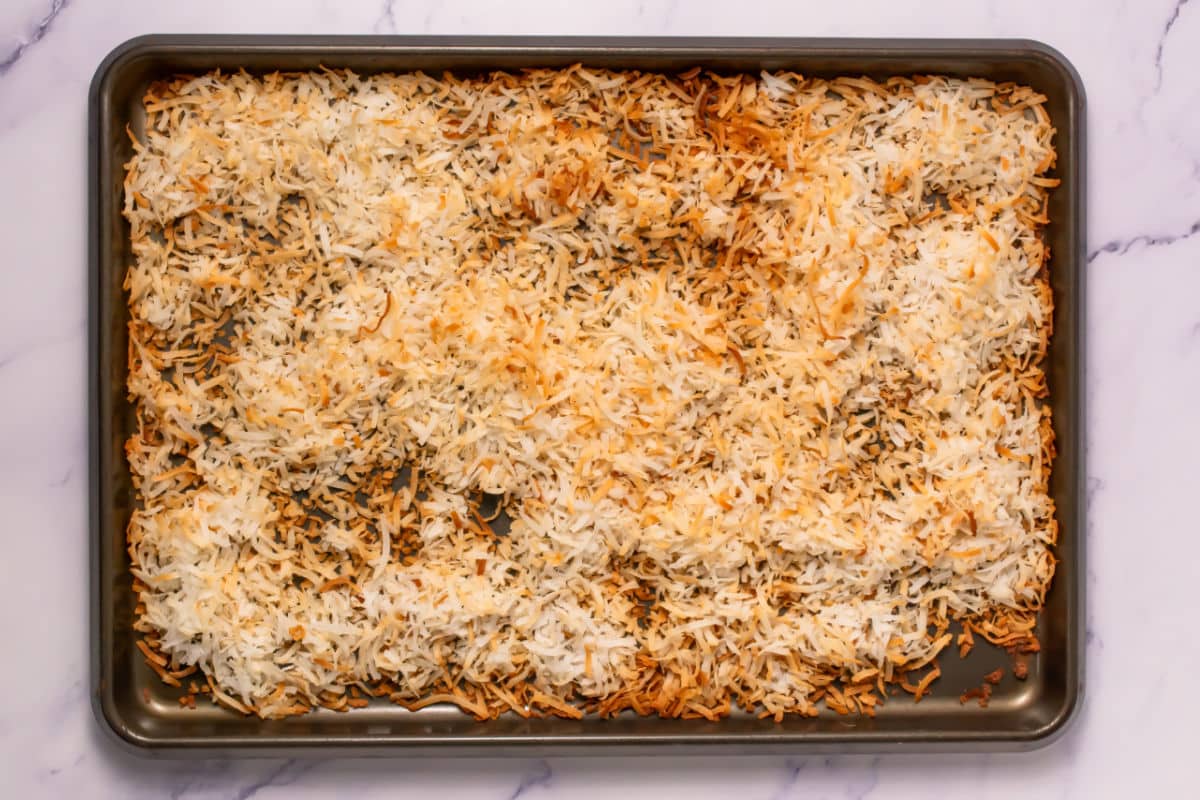 A baking sheet filled with shredded coconut, some toasted to a light golden brown for Somoa Copy Cat Cookies, on a marble surface.
