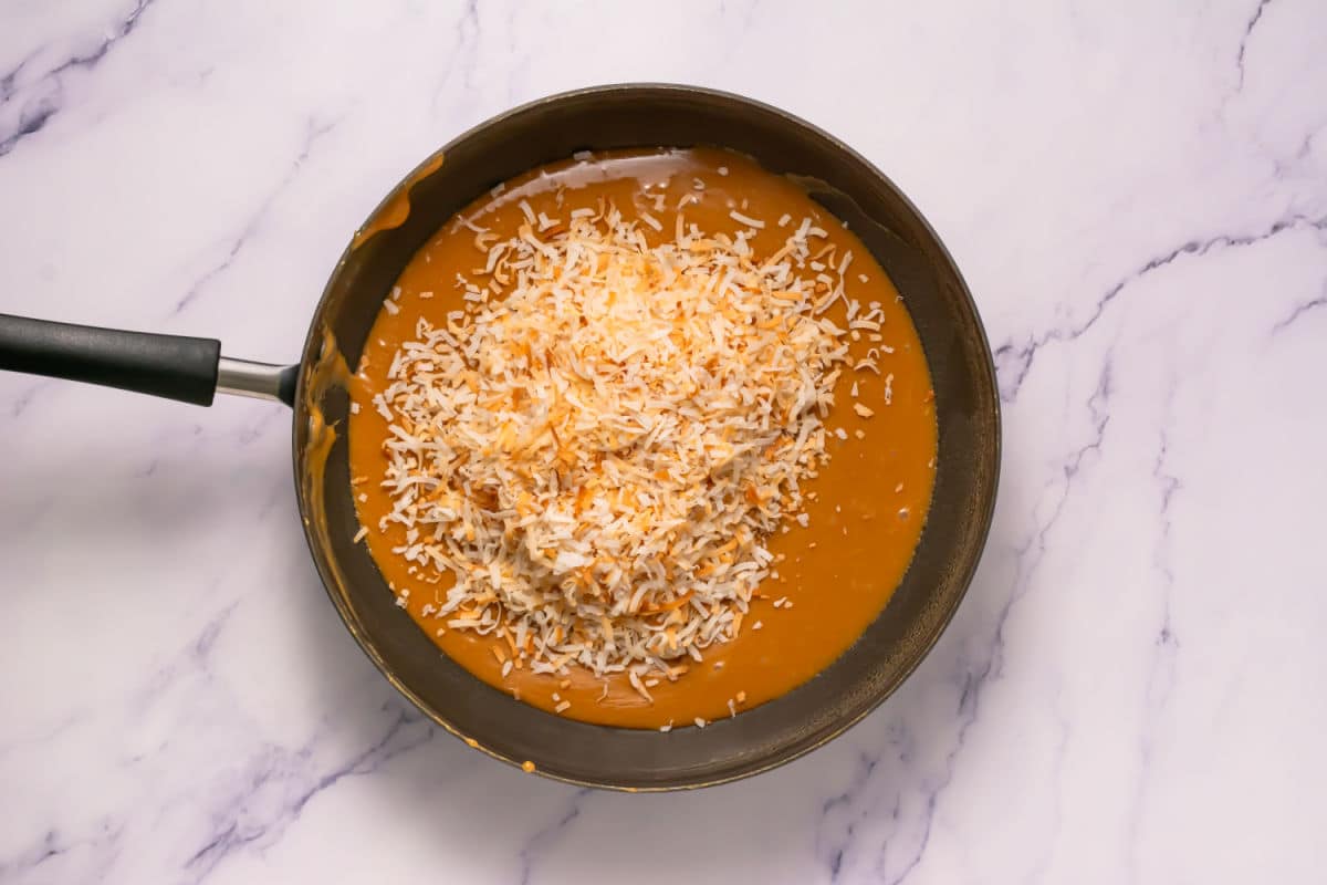 A skillet filled with caramel sauce topped with a generous layer of toasted shredded coconut, reminiscent of Somoa Copy Cat Cookies, set on a white marble surface.