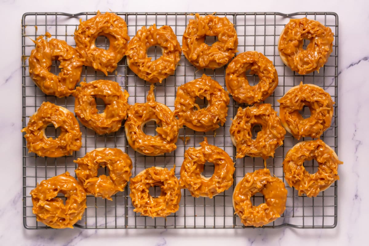 A cooling rack holds several Somoa Copy Cat Cookies, their caramel-coated, ring-shaped, and textured surfaces arranged on a white marble surface.